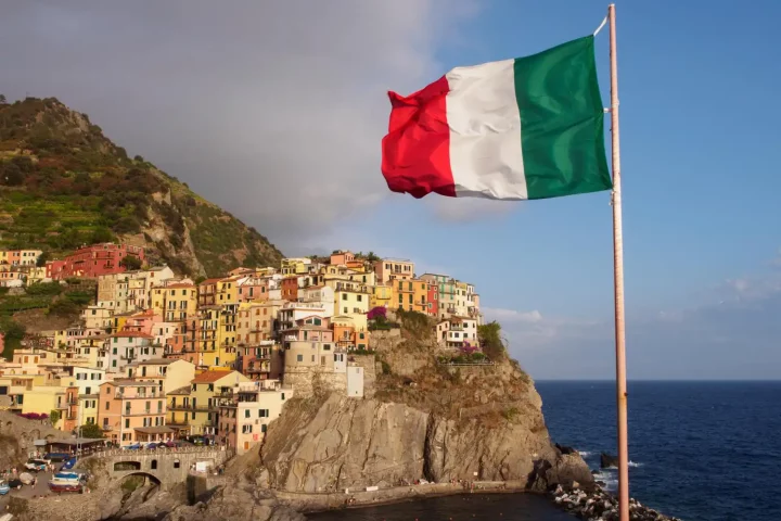 italian-regional-dialects The history of Italian regional dialects: Photo of the Italian flag in front of a cliffside village in Sicily